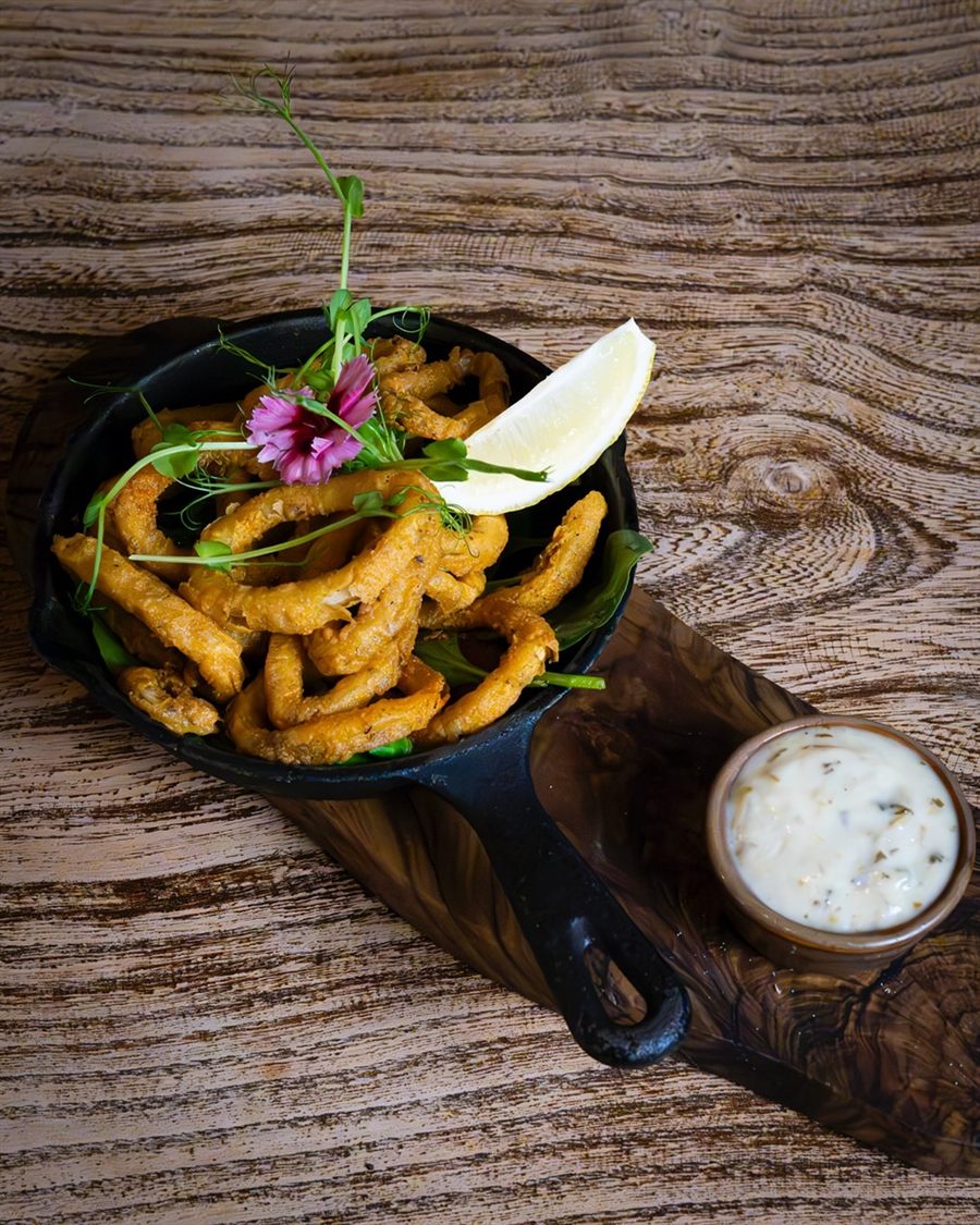 A plate of golden fried calamari rings with lemon wedges and a pot of tartare sauce.