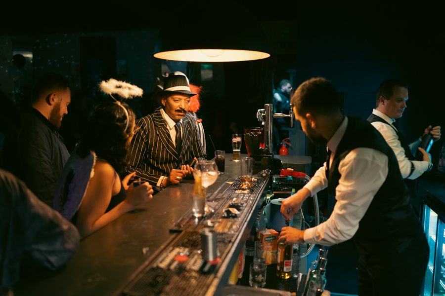 Guests in fancy dress chatting across the bar while staff mix drinks in the warm low light.