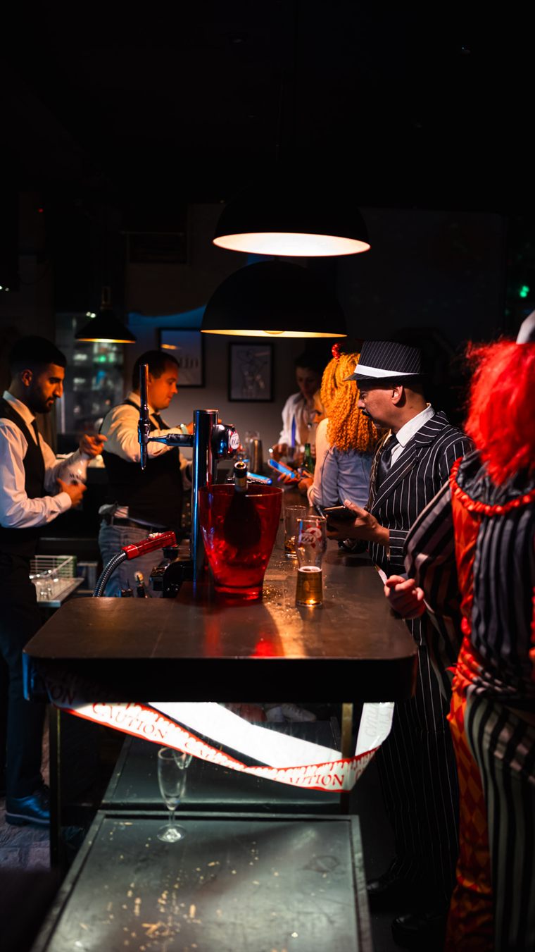 A busy party night: staff serving a crowd of guests in fancy dress, a red ice bucket on the bar top.