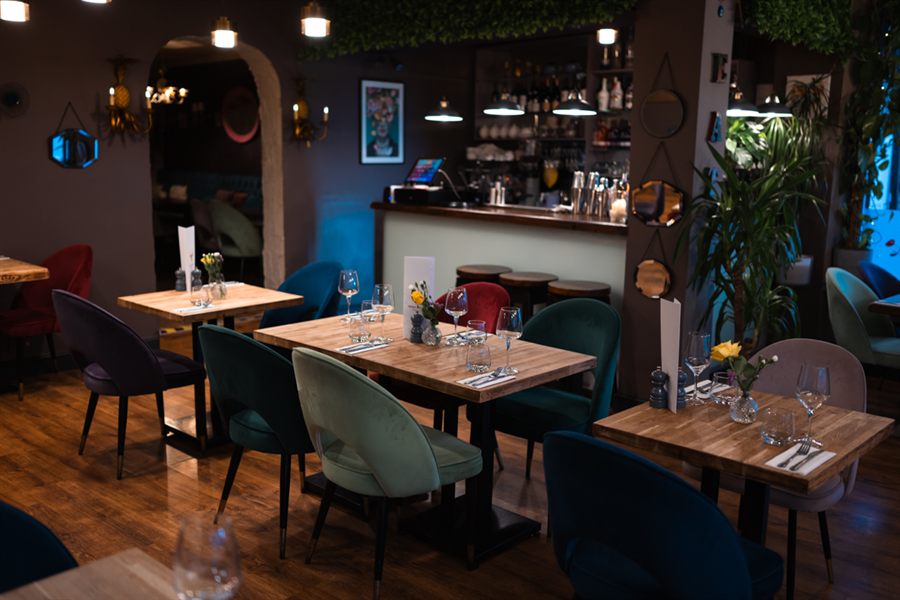 The dining room looking towards the bar: oak tables set with wine glasses, velvet chairs in green, teal and burgundy, and a lit back bar under pendant lamps.