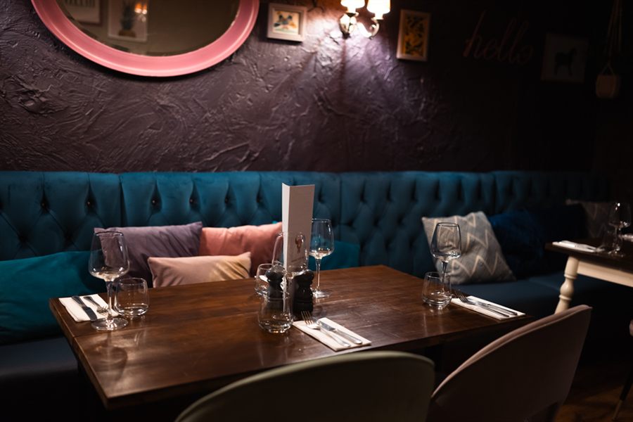 A table for four against a deep teal buttoned banquette scattered with cushions, under a pink-framed round mirror.