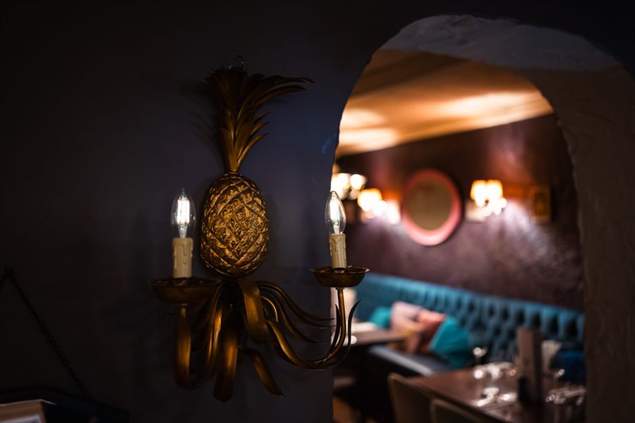 A gilded pineapple wall light with candle bulbs, and beyond it an archway through to the teal banquette in the next room.