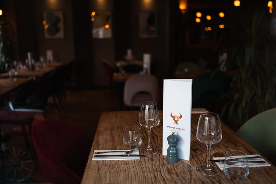 A close view of a laid oak table: wine glasses, folded napkin and cutlery beside the Mosaic Kitchen wine list.