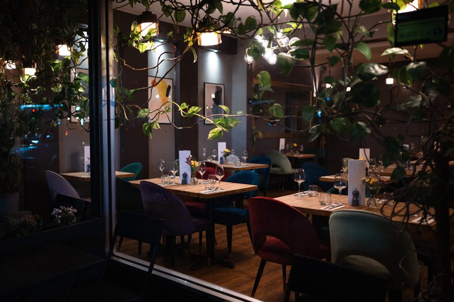The restaurant seen from the pavement at dusk through trailing greenery, warm lamplight over set tables inside.