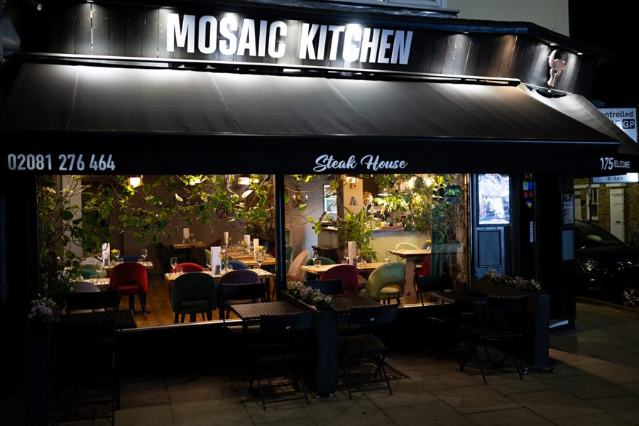 The Mosaic Kitchen shopfront at night: the lit sign and black awning over pavement tables, with the warm dining room glowing behind the glass.
